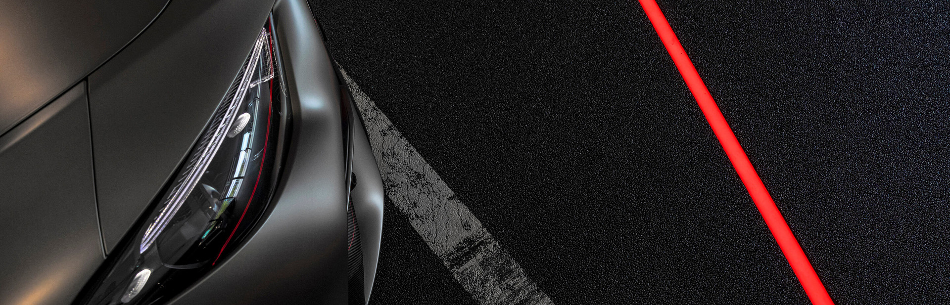 Ausschnitt eines eleganten Autos mit fokussierten Scheinwerfern, geparkt auf einem glitzernden schwarzen Untergrund mit einer kontrastreichen roten Linie.